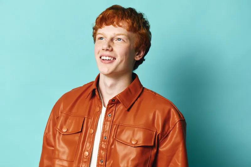 Young man with red hair wearing orange leather jacket smiling and looking away on blue background. Portrait of cheerful casual male with light skin and trendy outfit in studio.