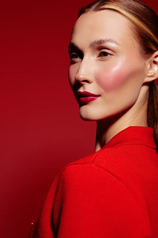 Close-up portrait of a woman with glowing skin and red lipstick wearing a red top against a red background. Concept of beauty, makeup, fashion, and elegance in studio lighting.