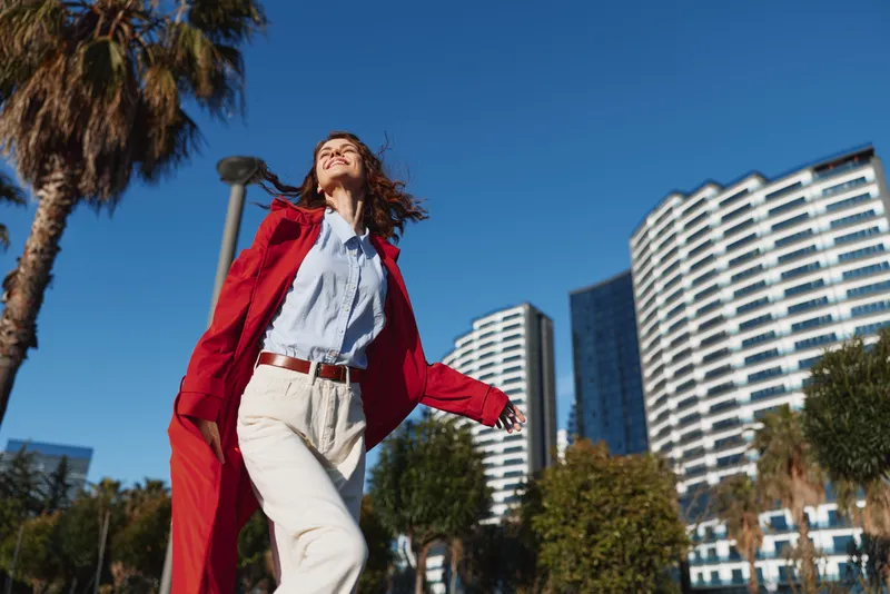 Confident woman in a red coat walks outdoors in a sunny city setting, with modern buildings and blue sky behind. A stylish urban moment of motion.