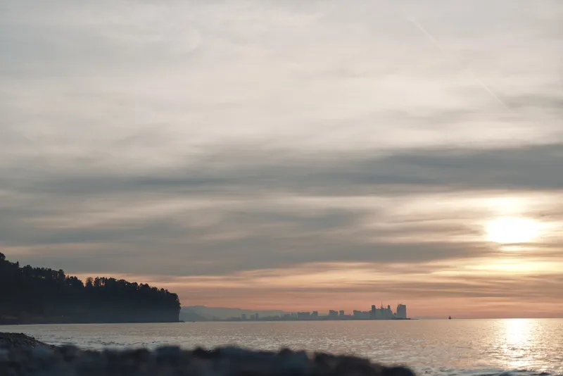 Sunset over calm sea with distant silhouettes along the horizon, a rocky shore in the foreground and soft clouds reflecting warm light across the water. creating a tranquil mood.