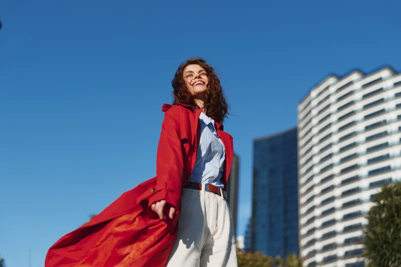 Young woman with curly hair wearing a red coat and white pants smiles confidently against a clear blue sky and modern buildings in the background.