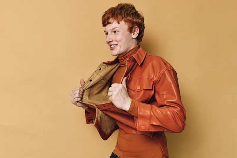 Young man with red hair wearing brown leather jacket over orange turtleneck sweater posing confidently on plain beige background indoors for fashion portrait style shoot.