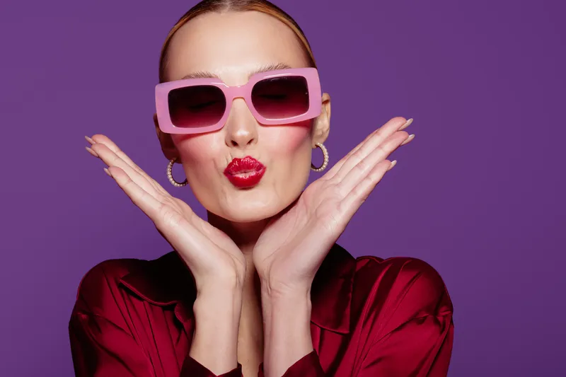 Stylish woman with pink sunglasses and red lipstick poses with hands near face on purple background. Fashion concept with bold makeup and confident expression in studio lighting.