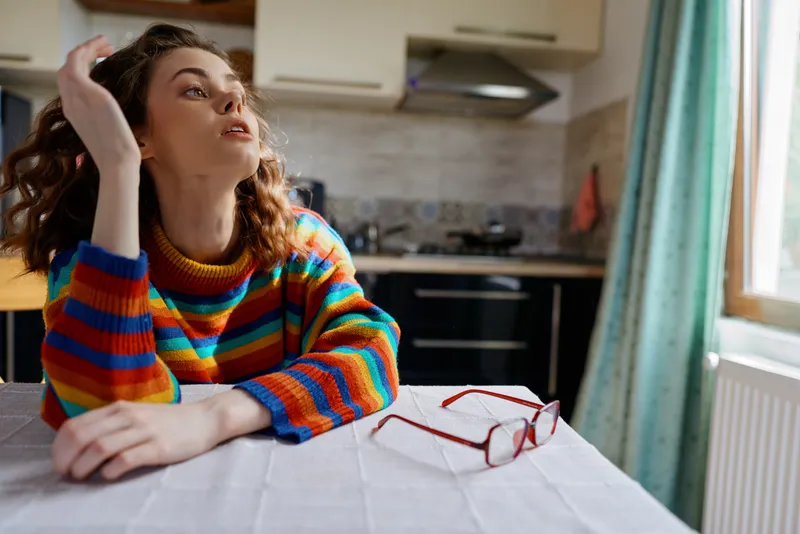 Young woman in a colorful striped sweater rests her chin on her hand at a kitchen table, gazing thoughtfully as daylight surrounds a cozy home setting.