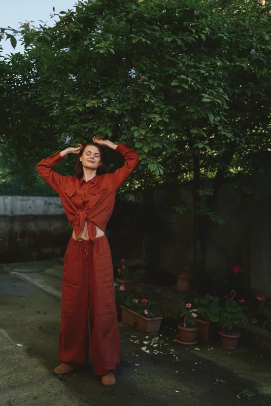 Woman in red wide-leg outfit posing with arms raised in a garden setting. Casual lifestyle scene with natural light, lush greenery and potted plants creating a warm, relaxed mood