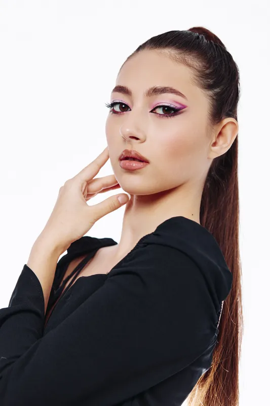 Young woman with artistic pink eye makeup and straight ponytail poses confidently in a black top on a white background. Concept for beauty creative fashion.