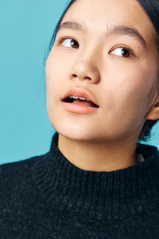 Young woman with open mouth and thoughtful expression looking sideways on light blue background, wearing black sweater, natural skin texture with visible pores and soft lighting on face.