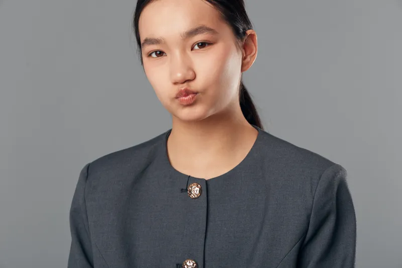 Portrait of young woman in gray jacket with buttons making kiss face against gray background. Female model with natural skin and dark hair in studio lighting for beauty concept.
