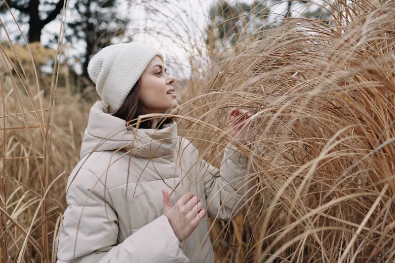 woman winter coat beanie grass field outdoor nature, serene portrait of a thoughtful model in soft neutral tones, capturing quiet solitude among tall grasses during a chilly day, exploration of calm