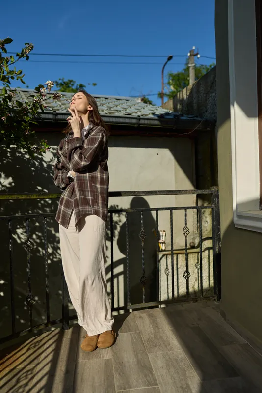 Young woman on a sunlit balcony wearing a plaid shirt and white pants, posing with her hand near her chin and savoring a calm morning mood as a soft breeze drifts