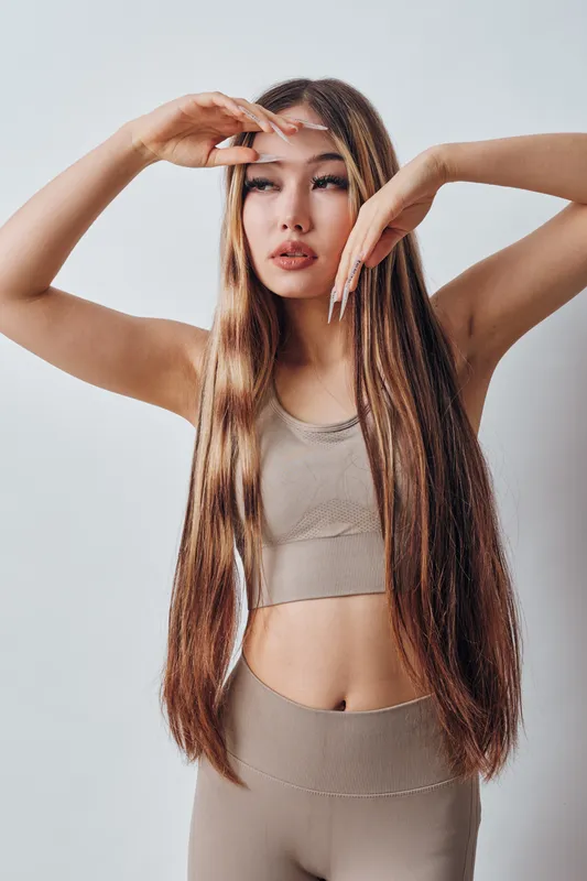Young woman with long straight hair wearing beige sportswear posing with arms raised against plain background. Fitness model, casual style, healthy lifestyle, confident look and natural beauty.