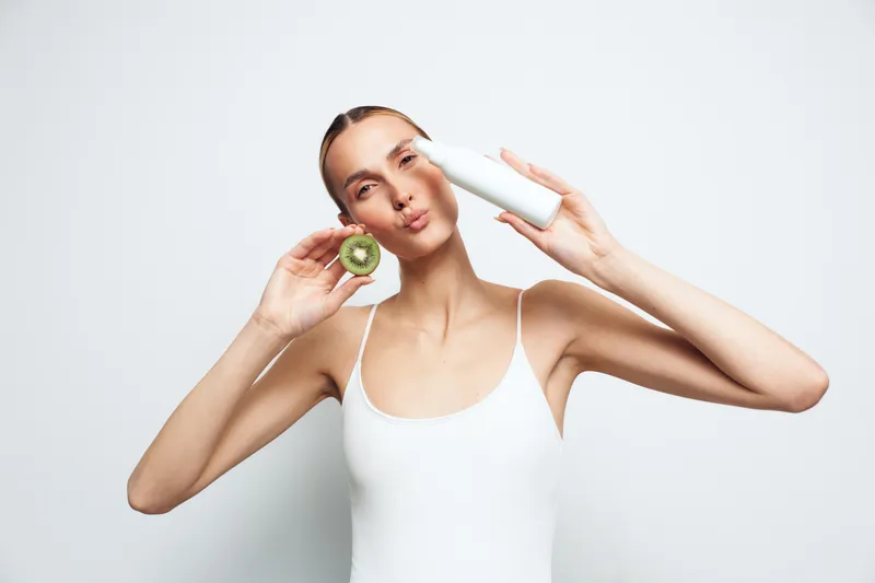 Young woman in white tank top holding kiwi slice near face and white bottle near eye with playful expression isolated on white background healthy lifestyle concept skincare beauty