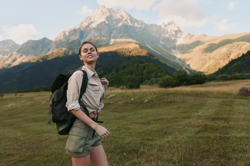 Young woman traveler with backpack hiking in mountains enjoys nature and fresh air during summer adventure with positive lifestyle and casual outfit.