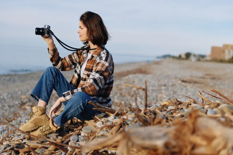 Photographer sits on a rocky beach with a camera, wearing a plaid shirt and jeans, capturing coastal scenery, driftwood and calm waves in a bright outdoor moment.