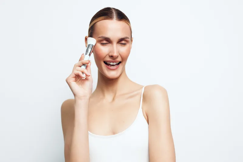 Young woman with closed eyes smiling applies makeup with brush on face isolated on white background, beauty routine concept, natural skin care and cosmetic application.