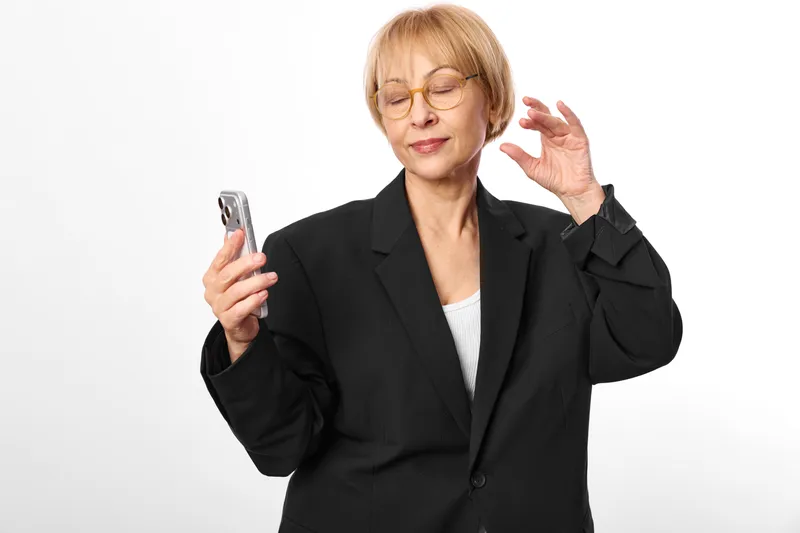 Elderly business woman in black blazer holds a smartphone with stressed expression, one hand raised. Glasses visible, isolated on white background, communication anxiety.