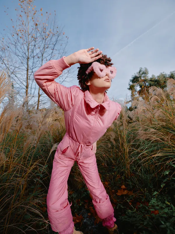 Pink jumpsuit model posing in a sunlit field, shielding eyes with a hand as the breeze moves through tall grasses, showcasing playful fashion and an outdoor lifestyle in nature.