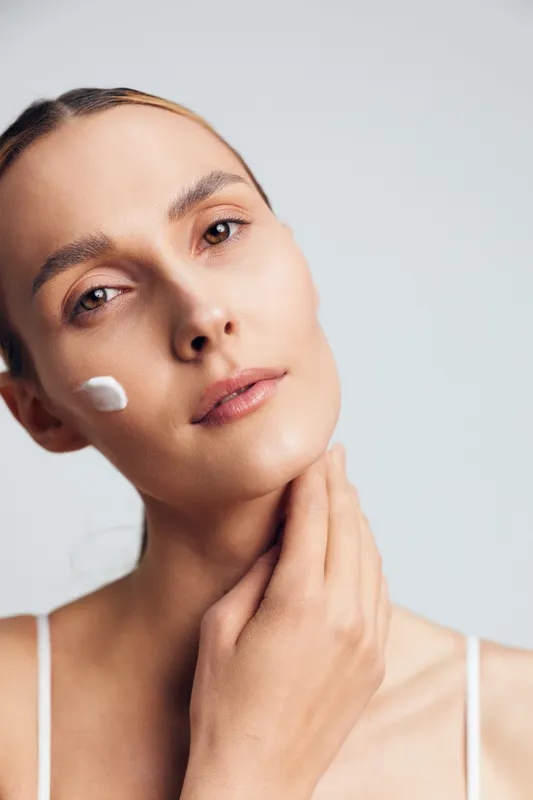 Young woman applies moisturizing cream on face with gentle hand touch, showing natural skin care routine, fresh healthy complexion, isolated on light background with soft lighting.