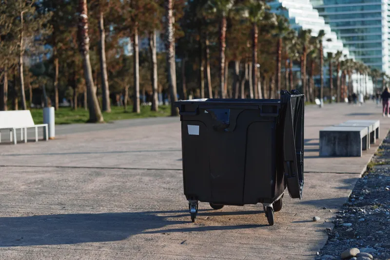 Black plastic trash bin with open lid standing on paved walkway near palm trees in urban park. Sunny day with clear sky and modern buildings in background.