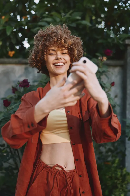 Young woman with curly hair takes a playful selfie in a sunlit garden, wearing a rust jacket and crop top, smiling at her phone