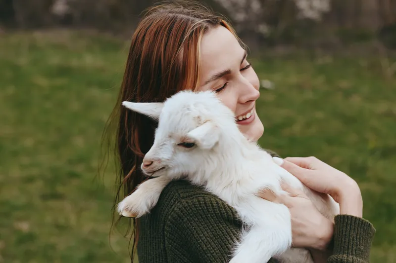 Joyful woman outdoors cradling a young goat kid, green meadow in the background, a warm, candid lifestyle moment that celebrates care, affection, and the bond between human and animal.
