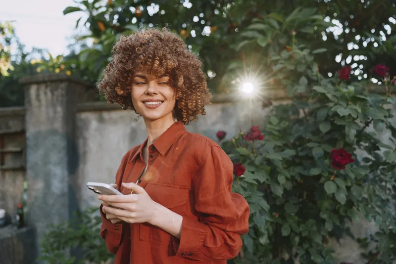Joyful woman enjoys an outdoor lifestyle moment, smiling while using a smartphone in a sunny garden, wearing a rust red blouse among flowers, capturing candid warmth and charm