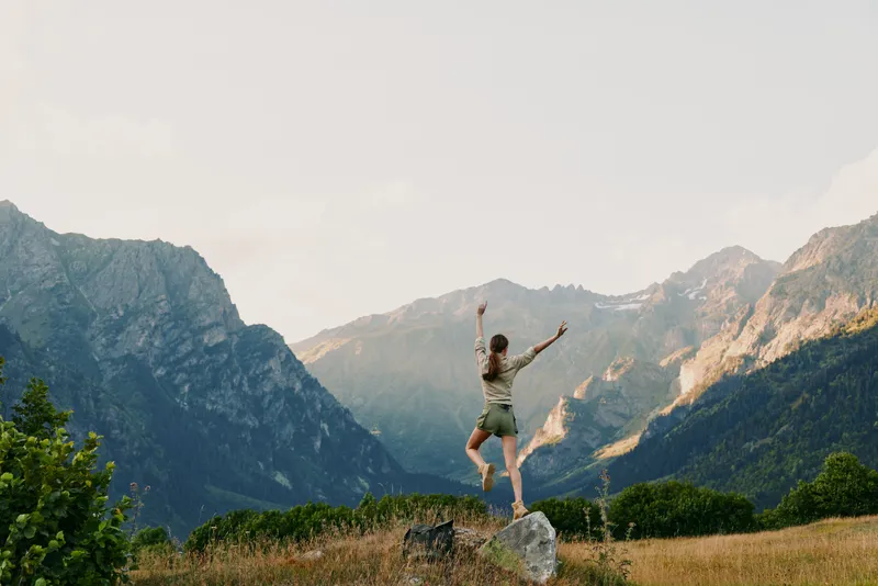 Young woman stands on a rocky outcrop with arms raised in celebration, framed by sunlit alpine valley and distant mountains, conveying freedom and a sense of adventure.