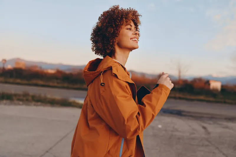 Curly haired woman in an orange jacket outdoors on a quiet road at sunset, portrait view. Active lifestyle, calm mood, natural light, soft wind, confident smile, roadside scenery.