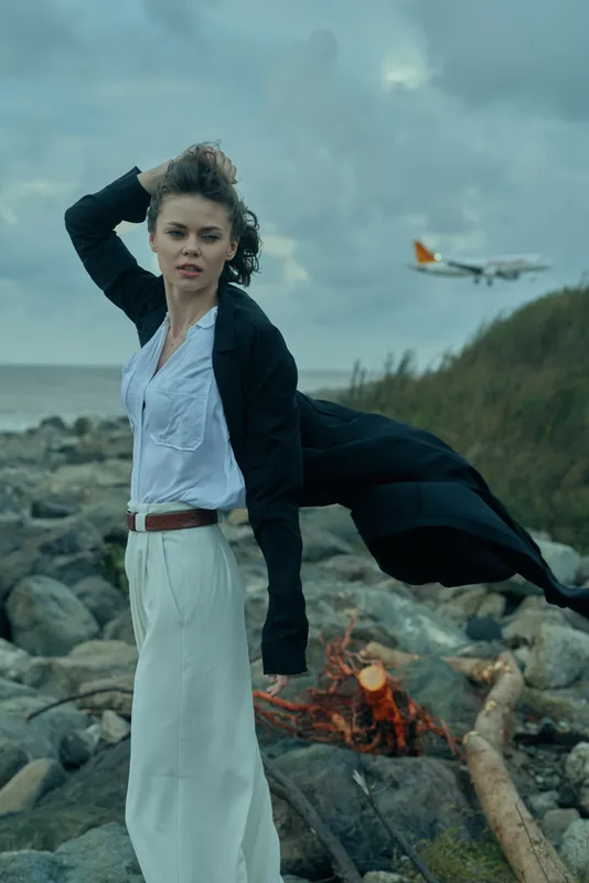 Wind, woman, coastline, fashion on a windy shore with flowing coat and white dress, ocean backdrop and distant aircraft in a moody sky, dramatic movement