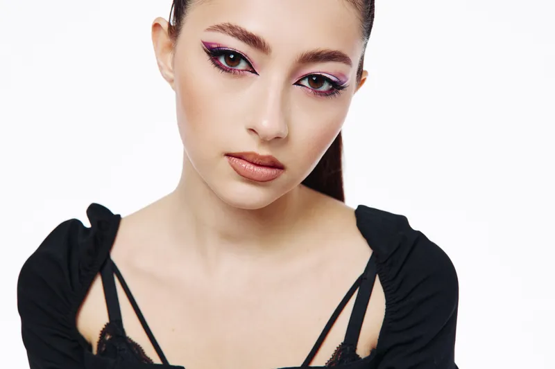 Close-up of a fashion woman with creative magenta eyeliner and natural lips wearing black off-shoulder top on white background. Concept of modern beauty and style portrait.