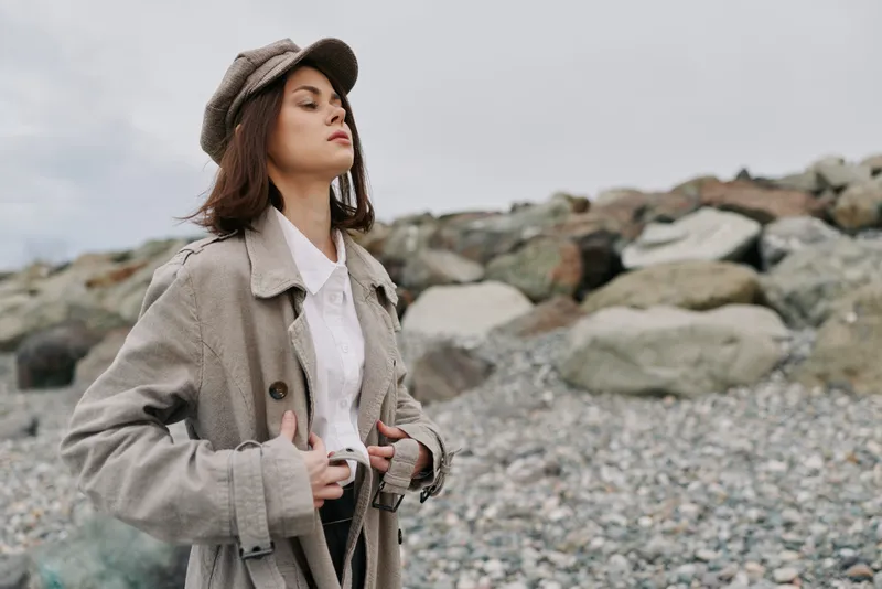 Young woman in vintage clothes stands on rocky beach with cloudy sky. She wears hat and coat, looking confident and serene. Concept of fashion, nature, and calm mood outdoors.