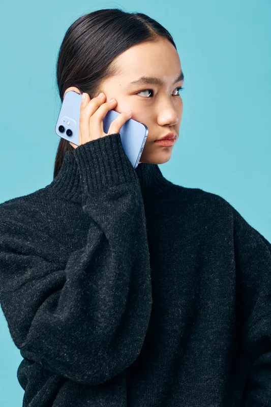 Woman talking on mobile phone wearing black turtleneck sweater against blue background. Casual communication, technology usage and portrait of young female with smartphone in hand.