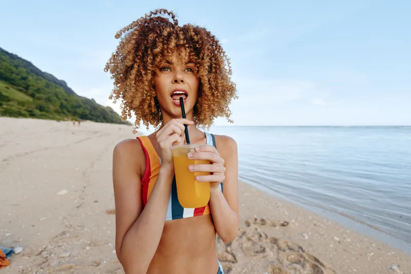 Woman with curly hair on a sunny beach holds a cup of juice with straw, looks contemplative and relaxed, summer vacation concept for hydration, healthy drink and refreshment outdoors today