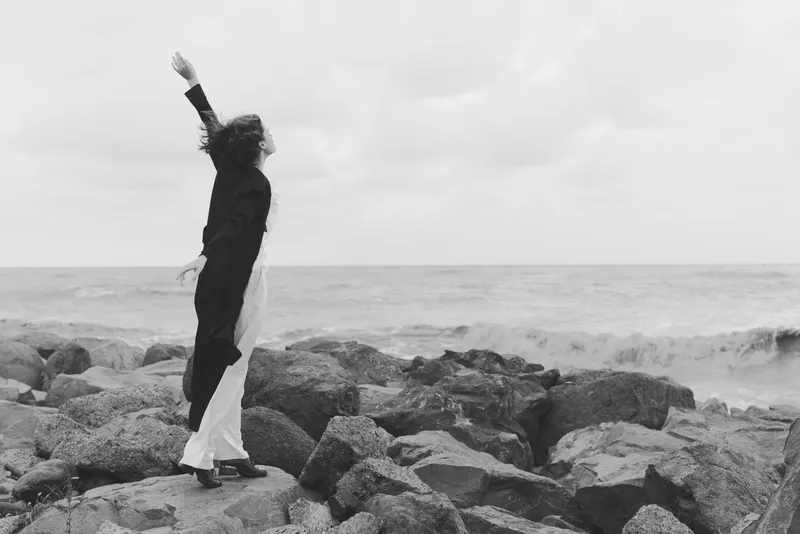 Woman stands on jagged rocks by the sea, arm raised against the wind in a dramatic monochrome scene, capturing solitude, resilience and a quiet moment of exploration along the rugged coastline