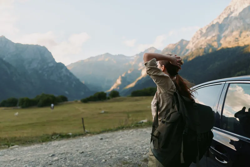 Woman hiking through a mountainous landscape with a backpack beside a parked car, embodying exploration, adventure, and outdoor freedom in wide open scenery.