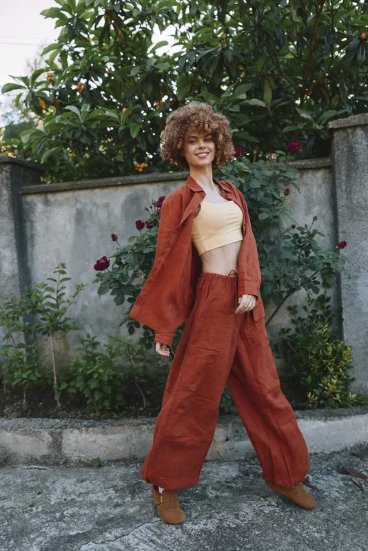 Confident woman in rust suit poses in a garden, showcasing contemporary lifestyle fashion, casual elegance, and natural joy in an outdoor setting with plants and a stone wall backdrop