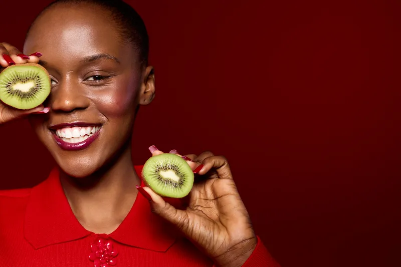Smiling black woman with dark skin holds two halves of fresh kiwi fruit near her face on red background. Concept of healthy lifestyle, natural food, and joyful mood with vibrant colors.