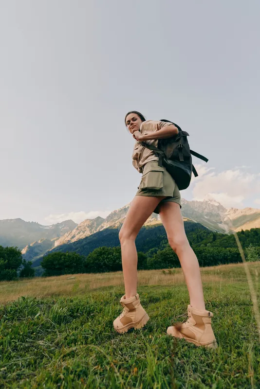 Young woman hiking uphill in a grassy meadow with distant mountains, wearing a backpack and beige shorts, looking determined and adventurous on a sunny day