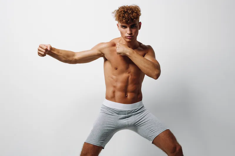 Young athletic man with curly hair practicing boxing stance shirtless in gray shorts on white background for fitness and martial arts training.