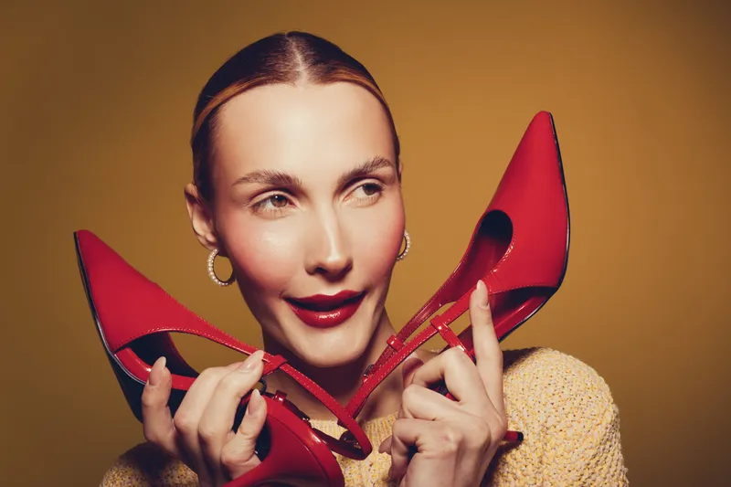 Young woman with red lipstick holds red high heels near face, wearing yellow sweater. Studio portrait on brown background. Concept of fashion, style, and beauty accessories.