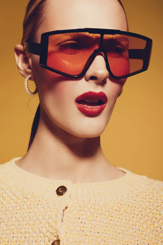 Young woman with oversized orange sunglasses and red lipstick poses against yellow background. Fashion portrait with trendy accessories and stylish knitted sweater. Concept of modern style.