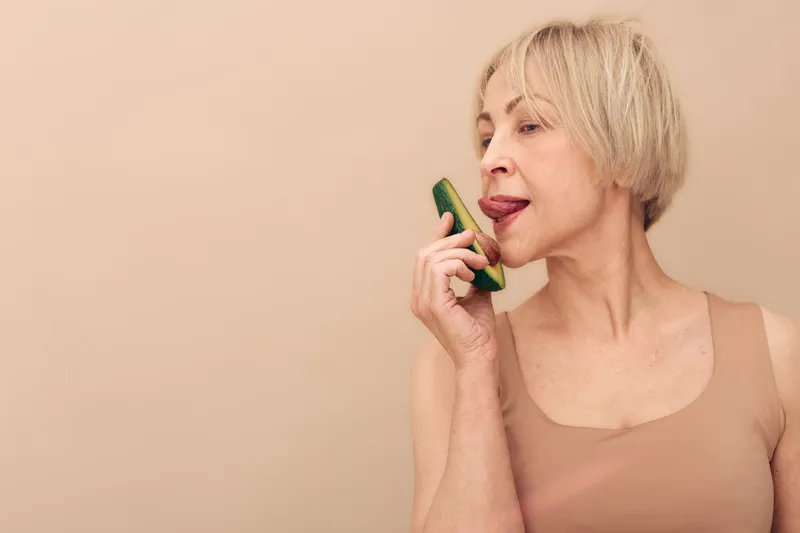 Blonde woman in beige tank top licking a fresh cucumber slice, isolated on beige background. Concept of healthy eating, natural skincare, and self-care lifestyle with fresh vegetable.