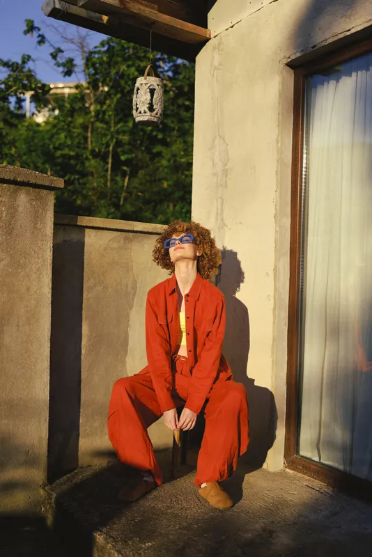 Woman in a bold red suit sits on a terrace wall, squatting pose, sunny outdoor setting, fashion forward vibe, casual elegance, warm light, architectural backdrop, lifestyle photography