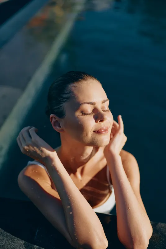 Relaxed woman in a swimsuit sits at the pool edge with eyes closed, warm sun on her skin, water droplets on her face, peaceful outdoor scene, natural colors, serene moment