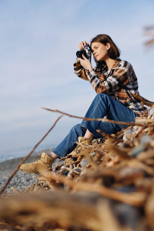 Female photographer outdoors by the coast, using a camera to capture sea scenery. Casual plaid shirt, jeans, and rocky foreground create a focused creative mood.