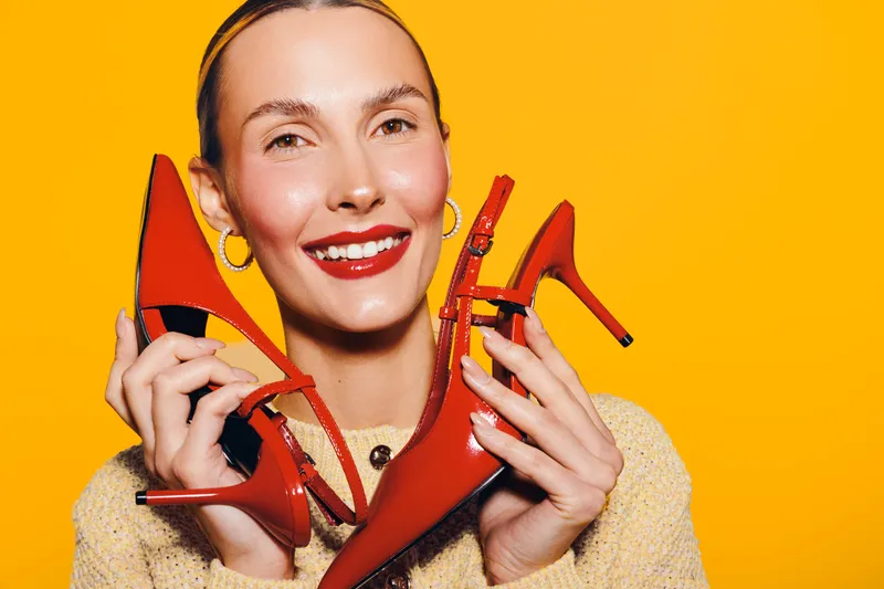 Smiling woman holds red high heel shoes near face on bright yellow background. Fashion concept with vibrant colors, beauty, and stylish accessories for footwear promotion or shopping.