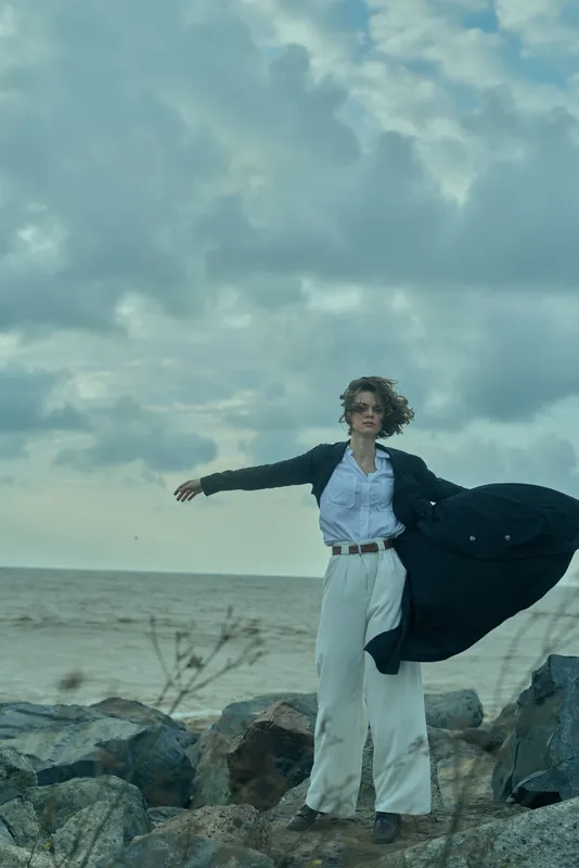 Fashionable woman standing on rocks by the sea, coat billowing in wind, white pants and blue shirt, moody clouds and dynamic coastal scenery, stylish and alive