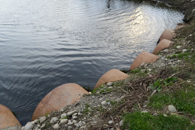 River with calm water by natural shoreline, stones and grass on the bank, sunlight glints on the surface, outdoor nature landscape with reflection and texture, peaceful lifestyle mood, quiet rural