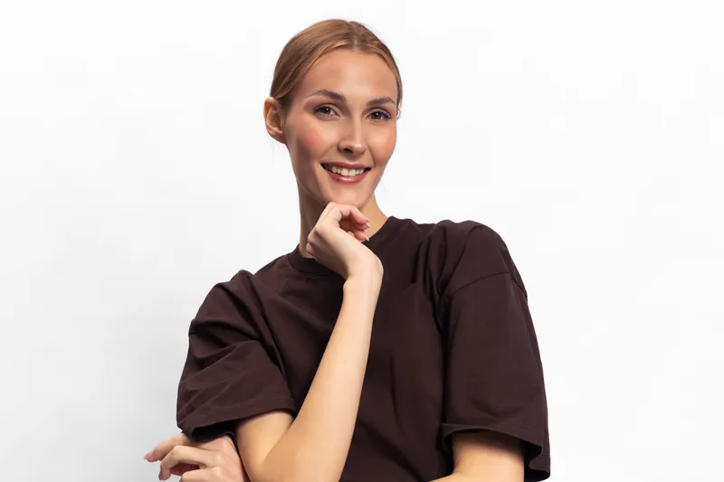 Smiling young woman with light hair wears casual brown t shirt, posing with hand near face against white background. Concept of happiness, confidence, and natural beauty in studio.