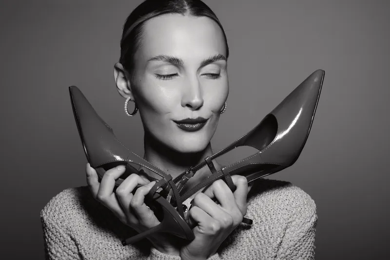 Elegant white woman with light skin holds two shiny high heels near her face with closed eyes, wearing knitted sweater and hoop earrings. Concept of fashion, style, and beauty.